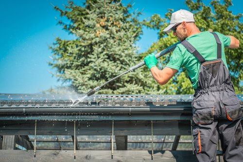 Male worker in a green shirt and gray overalls spraying down a house gutter with a pressure power washer.